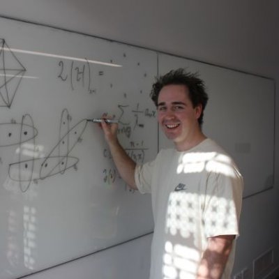 A man stands at a whiteboard, writing a mathematical equation. He is smiling at the camera.